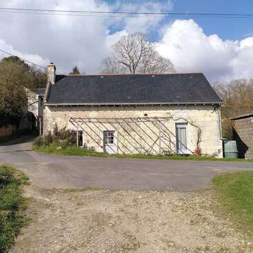 A rustic stone house with a dark roof sits by a quiet road, surrounded by trees and greenery, under a partly cloudy blue sky.