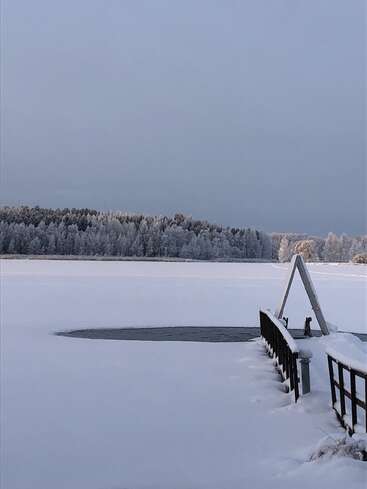 The image depicts a serene winter scene featuring a frozen lake, a snow-covered dock, and a forest of trees in the background, all blanketed in a layer of snow.