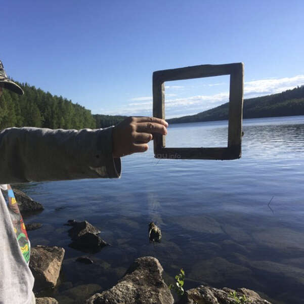A person holds a square frame in front of a serene lake, with rocks and trees in the foreground, set against a clear blue sky and distant hills.