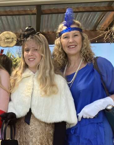 Two women dressed in vintage, 1920s-inspired attire smile together. One wears a blue dress, pearls, gloves, feathered headband; the other, a fur cape and small hat.