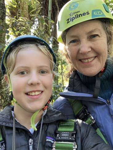 A smiling child and adult wearing helmets and harnesses, ready for an outdoor adventure at Rotorua Ziplines, surrounded by lush green forest scenery.