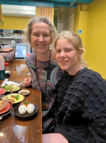 A smiling woman and girl sit together at a restaurant counter with various dishes of food, including cute animal-shaped buns, enjoying a cozy meal together.