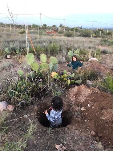 Dos niños juegan al aire libre entre cactus en un campo cubierto de hierba. Un niño está sentado en un gran agujero, el otro está sentado cerca, en el suelo.