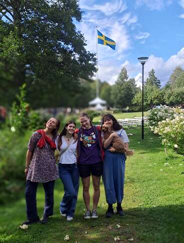 Cuatro personas sonrientes posan juntas sobre un frondoso césped verde, con un perro en brazos. Una bandera sueca ondea en lo alto, rodeada de árboles, flores y un cielo azul brillante.