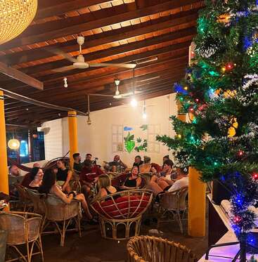 A lively group of people gather in a cozy, warmly lit room with wicker furniture and wooden ceiling. A decorated Christmas tree adds festive ambiance to the scene.