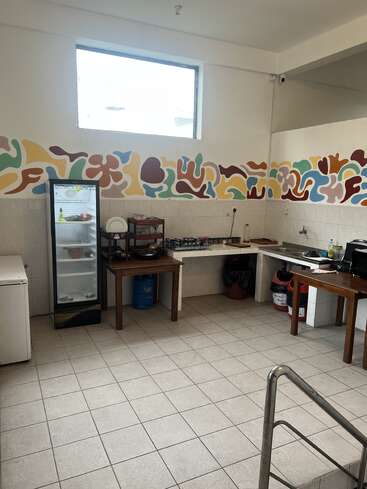 A simple kitchen with a tiled floor, colorful abstract wall art, refrigerator, sink, gas cylinder, dish rack, countertop, microwave, and large window providing natural light.