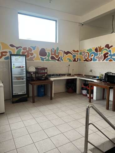 This kitchen features a refrigerator, tiled floors, colorful abstract wall art, a large window, sink, countertop, tables, and kitchen essentials, with a clean, airy atmosphere.