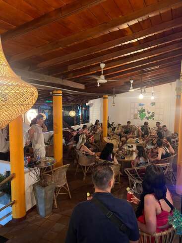 Un grupo de personas socializa en un salón acogedor y cálidamente iluminado con sillas de mimbre, pilares amarillos y bebidas, creando un ambiente relajado y animado en el interior.