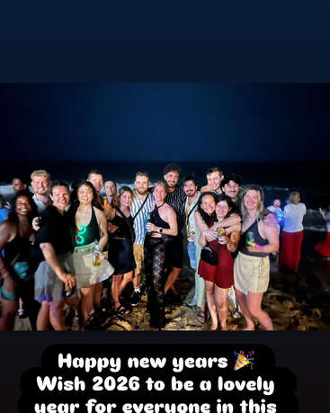 Um grupo alegre de amigos comemora a véspera de Ano Novo em uma praia à noite. Todos sorriem, segurando bebidas. A legenda deseja felicidades para 2026.