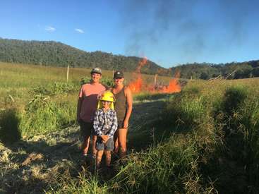 A imagem mostra uma família de três pessoas em um campo gramado, com um fogo controlado queimando ao fundo, em um céu azul claro.