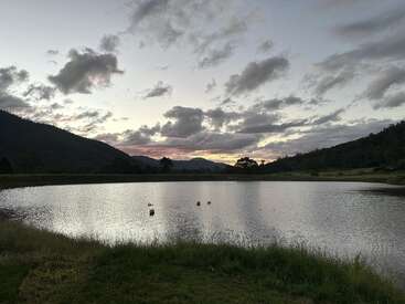 A imagem retrata uma cena serena de um lago ao pôr do sol, com uma margem gramada, montanhas distantes e um céu nublado, evocando uma sensação de paz e tranquilidade.