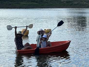 A imagem mostra três indivíduos em um pequeno barco laranja em um corpo de água sereno, cada um segurando um remo e usando chapéus, com um objeto amarelo visível ao fundo.