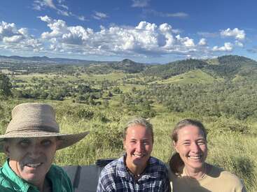 A imagem mostra três pessoas posando para uma selfie em um pitoresco campo gramado, com colinas e árvores visíveis ao fundo sob um céu azul com nuvens brancas.