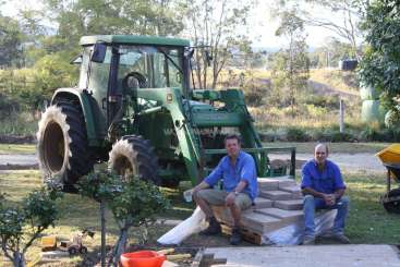 A imagem mostra dois homens sentados em uma pilha de madeira, com um trator verde ao fundo, cercado por árvores e uma estrada de terra.