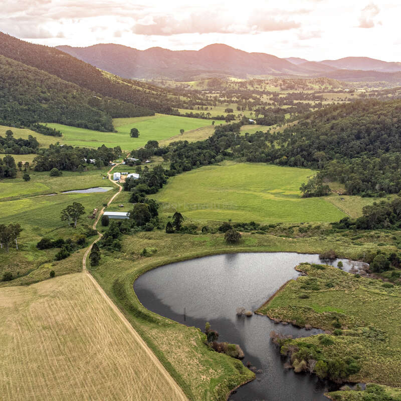 A imagem retrata uma paisagem rural serena com colinas onduladas, um rio sinuoso e um conjunto de edifícios, tendo como pano de fundo montanhas distantes sob um céu nublado.
