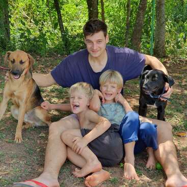 L'image représente un homme assis par terre avec deux jeunes garçons et deux chiens, entourés d'arbres et de verdure luxuriante dans un cadre extérieur serein.
