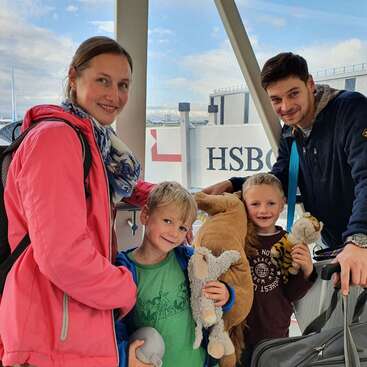 L'image montre une famille de quatre personnes posant pour une photo dans un aéroport, avec une valise et des animaux en peluche, sur fond de ciel nuageux et d'un grand panneau indiquant "HSBC".