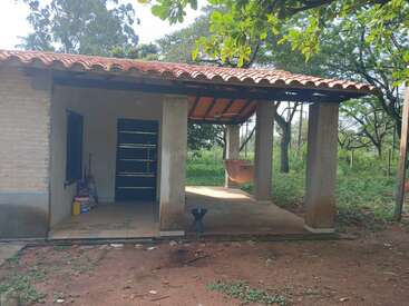 Une petite maison rurale avec un toit en tuiles et un patio ouvert se dresse parmi les arbres. Un hamac est suspendu, des produits d'entretien sont posés près de la porte. Un cadre paisible et rustique.