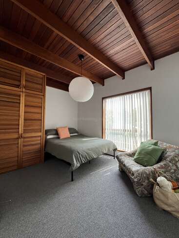 This cozy bedroom features a wooden ceiling, a bed with green bedding, a patterned couch, large window with vertical blinds, wooden closet, and soft lighting.