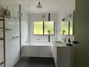 This bathroom features a modern design with a glass shower, bathtub, large frosted window, white tiles, black accents, plants, vanity, mirror, and minimalistic decor.