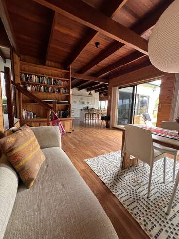 This cozy, open-plan living space features wooden ceilings and floors, a comfortable sofa, dining area, bookshelf, and a kitchen with abundant natural light from large windows.