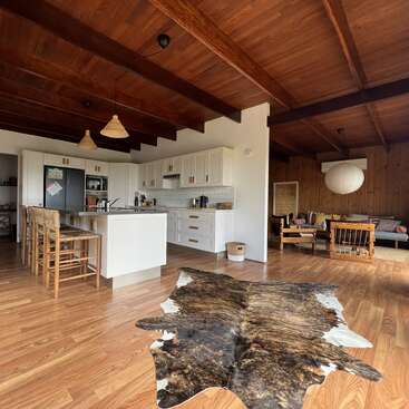 This image shows a modern, open-plan kitchen and living room with wooden floors, exposed beams, white cabinets, island seating, a cowhide rug, and cozy décor.