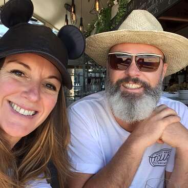 Una mujer sonriente con una gorra negra con orejas de Mickey Mouse se sienta junto a un hombre barbudo con gafas de sol y sombrero de paja, ambos disfrutando de un soleado día de café.