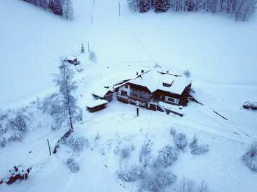 A large wooden house is surrounded by deep snow, trees, and a serene winter landscape. The view is aerial, capturing peaceful, isolated, frosty beauty.