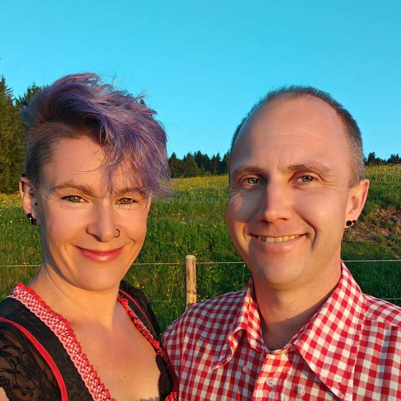 A smiling couple poses outdoors in warm sunlight. The woman has purple hair, a nose ring, and a checked dress. The man wears a red checked shirt.