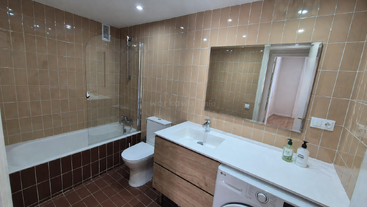 Cette salle de bains est dotée de murs carrelés marron, d'une baignoire avec une paroi en verre, de toilettes, d'un meuble-lavabo en bois avec un lavabo, d'un grand miroir et d'un lave-linge.
