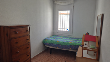 Il s'agit d'une petite chambre toute simple, avec un lit simple, un couvre-lit coloré, une commode en bois, une maison de poupées, une fenêtre à barreaux et des murs blancs unis.
