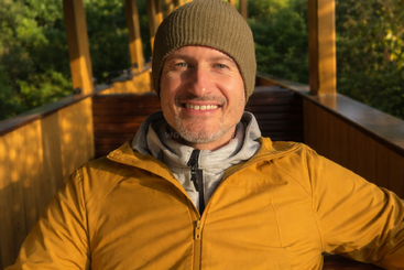A smiling man in a brown beanie and yellow jacket sits on a sunlit wooden porch, surrounded by lush green trees, enjoying a peaceful, pleasant morning.