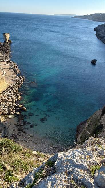 A breathtaking cliffside view overlooks the serene blue sea, with rocky formations along the shore, clear turquoise water, and distant coastline under a bright sky.