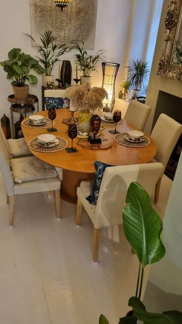 Este acogedor comedor cuenta con una mesa de madera para seis comensales, rodeada de sillas color crema, exuberantes plantas verdes, detalles decorativos, obras de arte en la pared e iluminación tenue.