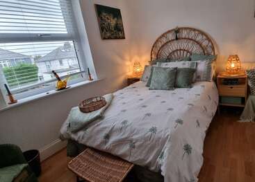 Un acogedor dormitorio con cabecero de ratán, ropa de cama verde tropical, cálidas lámparas de mesilla, cesta de mimbre, gran ventanal, paredes neutras, suelo de madera y acentos de decoración frondosa.