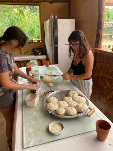 Two women are smiling and shaping dough in a cozy kitchen. There are round dough balls on a tray, surrounded by ingredients, cups, and kitchen utensils.