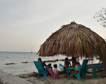 Una familia se relaja en sillas de colores bajo una sombrilla de paja junto a la playa. El mar en calma, las rocas dispersas y un velero lejano completan la escena.