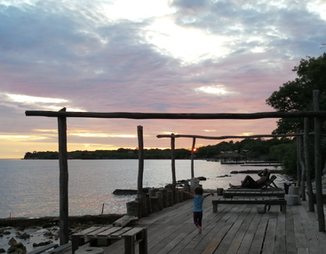Una apacible puesta de sol junto al agua, un muelle de madera con bancos, un niño paseando, un adulto relajándose, rodeados de árboles bajo un hermoso cielo de colores.