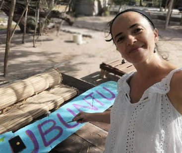 Una mujer sonriente vestida de blanco pinta la palabra "UBUNTU" en morado sobre una tabla de madera azul al aire libre, rodeada de bancos y árboles.