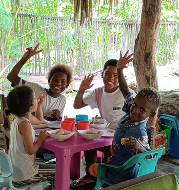 Cuatro niños sentados alrededor de una pequeña mesa rosa al aire libre, sonríen y saludan alegremente mientras disfrutan de una comida juntos. Una exuberante vegetación rodea la alegre escena, creando calidez.