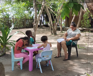 Tres niños se sientan en una mesita al aire libre, concentrados en dibujar o escribir, mientras un adulto se sienta cerca, en una silla, para ayudarles y supervisar su actividad.
