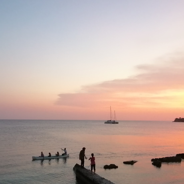 Una hermosa puesta de sol sobre las tranquilas aguas del océano, con piragüistas remando cerca, dos personas de pie sobre las rocas y un velero lejano que realza la tranquila escena.