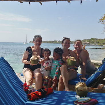 Tres adultos y dos niños se relajan en tumbonas azules junto al mar, disfrutando de cocos frescos. Día soleado, ambiente tropical, con barco y árboles al fondo.