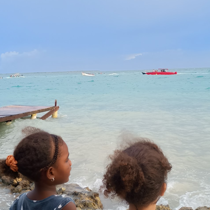 Dos chicas jóvenes están de pie en la orilla rocosa, contemplando el océano turquesa. Cerca hay un muelle de madera roto. Barcos, entre ellos uno rojo, flotan en aguas tranquilas.