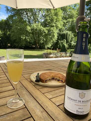 Una mesa de madera puesta al aire libre con una copa de vino espumoso, un pastel parcialmente comido y una botella de Crémant de Bourgogne, bajo una sombrilla, con vistas al jardín.