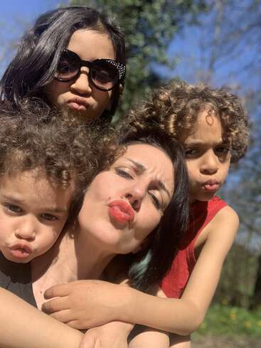 The image shows a woman posing with two children, both making kissy faces, in a sunny outdoor setting with trees and blue sky in the background.