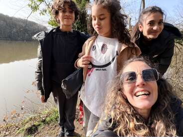 A woman and three children enjoy a sunny day outdoors by a lake. They smile and pose together for a cheerful selfie surrounded by nature.