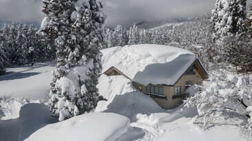 Uma casa grande está coberta de neve, com uma árvore grande à esquerda e uma menor à direita. A casa tem um telhado inclinado e uma chaminé. O fundo é um céu claro.