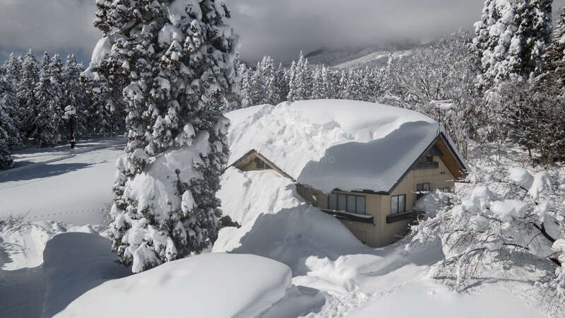Uma casa grande está coberta de neve, com uma árvore grande à esquerda e uma menor à direita. A casa tem um telhado inclinado e uma chaminé. O fundo é um céu claro.