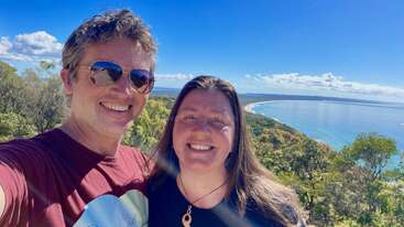 A smiling couple takes a selfie outdoors on a sunny day, with a scenic view of the coastline, blue sky, lush greenery, and ocean in the background.